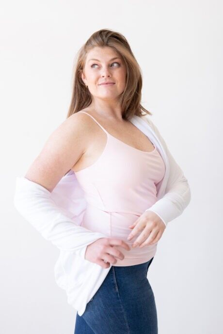 A woman wearing a pink tank top under a white cardigan, showcasing a scoop neck spaghetti strap layering camisole.